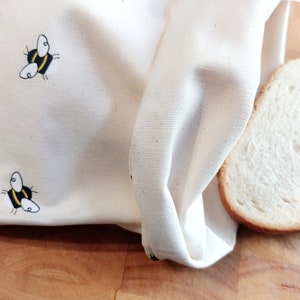 K&ouml;nnte beinhalten: Eine wei&szlig;e Brotbeutel aus Baumwolle mit einem Bienenmuster. Der Beutel ist &uuml;ber eine Scheibe Wei&szlig;brot auf einer Holzoberfl&auml;che gefaltet.