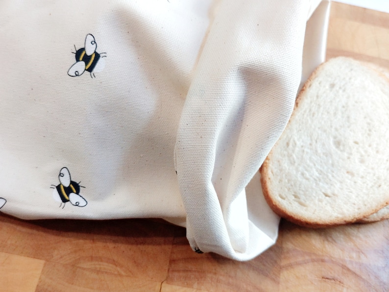 K&ouml;nnte beinhalten: Eine wei&szlig;e Brotbeutel aus Baumwolle mit einem Bienenmuster. Der Beutel ist &uuml;ber eine Scheibe Wei&szlig;brot auf einer Holzoberfl&auml;che gefaltet.