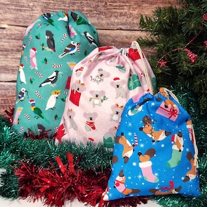 May include: Three fabric drawstring bags with holiday-themed prints. The large teal bag features various birds, the medium pink bag displays koalas, and the small blue bag shows platypuses, all accented with festive holiday elements.