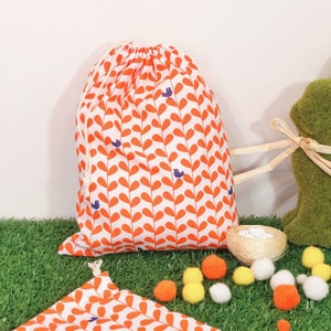 May include: A drawstring bag featuring a white background with an orange leaf pattern and small blue bird motifs. The bag is tied with a white cord. A matching fabric square is visible in the foreground. The bag is likely made of cotton.
