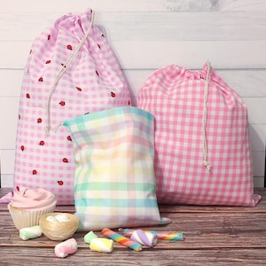 May include: Three drawstring bags are displayed. Two are pink gingham with ladybug patterns, and one is pastel plaid. A cupcake, sweets, and marshmallows are in front of the bags. The bags are on a wooden surface.