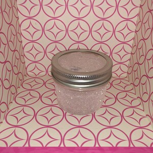 Op de afbeelding: Een glazen pot met een zilveren deksel is gevuld met witte glitter. De pot staat op een wit en roze gepatroneerd oppervlak.