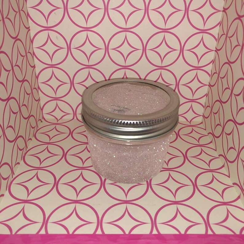 Op de afbeelding: Een glazen pot met een zilveren deksel is gevuld met witte glitter. De pot staat op een wit en roze gepatroneerd oppervlak.