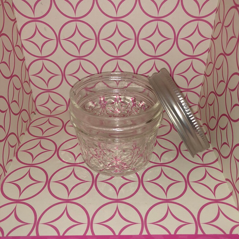 Op de afbeelding: Een glazen pot met een zilveren deksel. De pot heeft een getextureerd oppervlak en staat op een roze en witte gepatroneerde achtergrond.
