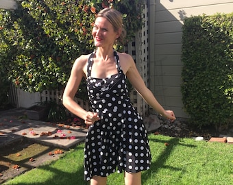 May include: A woman wearing a black and white polka dot dress with a halter neckline. She is standing on a grassy lawn.