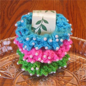 May include: Stack of three crocheted scrunchies in blue, pink, and green, each adorned with white pearl beads. A decorative tag with a leaf design is nestled in the center. The scrunchies are displayed on a clear, patterned glass plate.