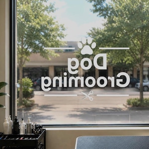 以下が含まれることがあります： 白いグラフィックで「Dog Grooming」と鏡文字で書かれた窓。足跡とハサミが描かれています。窓からは、木や建物のある通りが見えます。