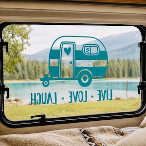 May include: A teal vinyl decal applied to a window, featuring a graphic illustration of a vintage camper trailer with a heart shape on the door. Below the camper, the text reads LIVE - LOVE - LAUGH in a bold, sans-serif font style.