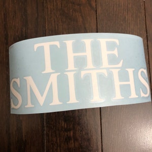 May include: A light blue vinyl decal featuring the white text "THE SMITHS" in uppercase. The decal is curved, suggesting it's ready for application. The lettering is in a clear, traditional font, suitable for various surfaces.