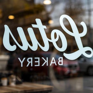 May include: A storefront window with the word "Lola's" in a cursive font, above the word "Bakery" in a sans-serif font, both in white. The reflection shows cars and a table.