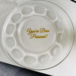 Può includere: Un piatto in vetro trasparente caratterizzato da una serie di rientranze circolari lungo il bordo e dal testo "You've Been Poisoned!" stampato in lettere dorate al centro. Il design è circolare con una superficie liscia e trasparente.