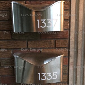 May include: Two brushed metal mailboxes fixed to a brick wall. The top mailbox has the number 1337 in white, the bottom one displays 1335. The mailboxes have a curved design and a contemporary look. The wall is made of red bricks.
