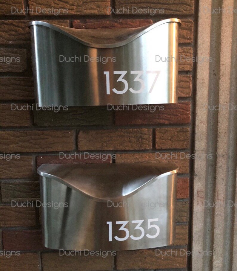 May include: Two brushed metal mailboxes fixed to a brick wall. The top mailbox has the number 1337 in white, the bottom one displays 1335. The mailboxes have a curved design and a contemporary look. The wall is made of red bricks.