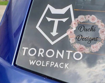 May include: A white vinyl decal applied to a blue vehicle window featuring the Toronto Wolfpack logo, the text TORONTO WOLFPACK, and a circular floral wreath graphic with the script text Duchi Designs in the center.