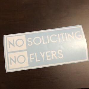 May include: A blue and white sticker with the text "NO SOLICITING NO FLYERS" in white lettering.