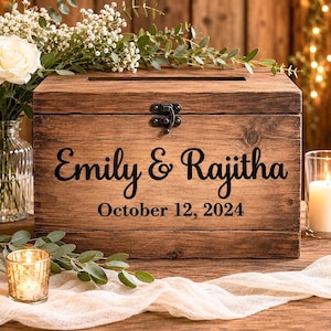 May include: A wooden card box with a slot and latch. It's engraved with "Emily & Rajitha" and "October 12, 2024". The box is surrounded by flowers and candles, creating a warm, rustic aesthetic. The box is made of wood.