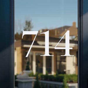 May include: White house number "714" displayed on a glass door. The numbers are in a serif font, contrasting against the blurred background of a house and landscaping. The door is framed by a dark, possibly black, door frame.