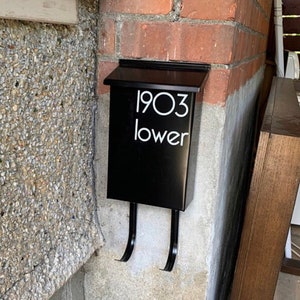 May include: A black mailbox fixed to a brick wall. The mailbox features a hinged lid and two curved arms. White text on the front reads "1903 lower". The mailbox is mounted on a concrete and brick wall.