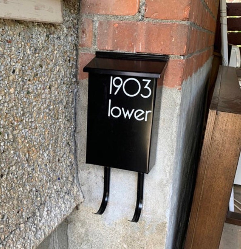May include: A black mailbox fixed to a brick wall. The mailbox features a hinged lid and two curved arms. White text on the front reads "1903 lower". The mailbox is mounted on a concrete and brick wall.