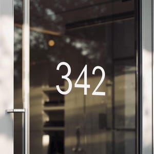 May include: A glass door features the white numerals "342". The numbers are in a modern, sans-serif font. The door has a silver handle and frame, reflecting the surroundings. The overall aesthetic is clean and contemporary.