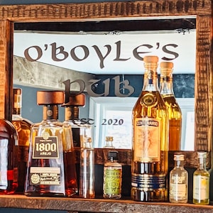 Puede incluir: Espejo enmarcado en madera con las palabras "O'Boyle's Pub". La repisa inferior contiene varias botellas de licor, incluyendo tequila y ginebra. El espejo refleja una ventana y el año "2020".