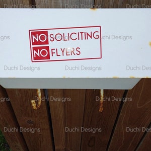 May include: White metal sign with red lettering that says "NO SOLICITING" and "NO FLYERS". The sign is mounted on a wooden fence.