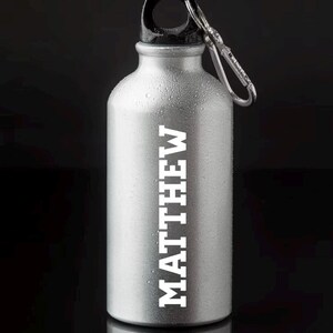 Op de afbeelding: Een zilveren waterfles met een zwarte dop en een draagriem. De fles heeft de naam "MATTHEW" in wit erop gedrukt.