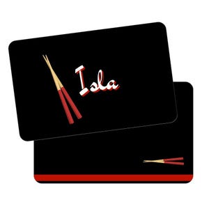 May include: Black placemat with rounded corners, featuring the name "Isla" in white with a red outline. A pair of red and tan chopsticks is on the top placemat. A second placemat is visible below, with a red stripe and chopsticks.