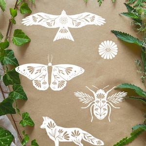 Peut inclure: Découpages en papier blanc d'un oiseau, d'un papillon, d'un coléoptère, d'un renard et d'une fleur sur fond marron. L'oiseau et le papillon ont des motifs floraux. Le feuillage vert et un tissu gris encadrent la scène.