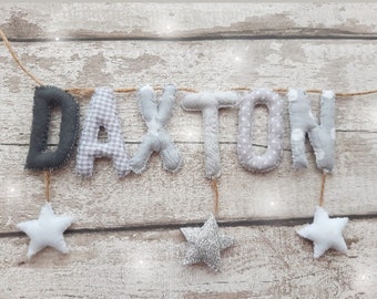 Op de afbeelding: Een banner met de naam "DAXTON" in grijs en witte stoffen letters. De letters zijn geregen aan een natuurlijke touw, met witte en zilveren sterren als accenten. De achtergrond is een licht houten patroon.