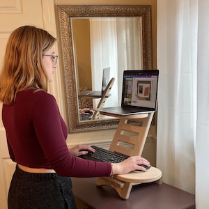 May include: A light-colored wooden laptop stand with a keyboard and mouse. The stand has a tiered design with a circular base and a rectangular top for the laptop. The stand is on a dark surface.