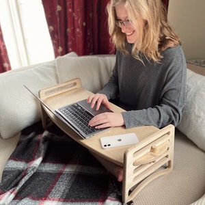 Large Adjustable lap desk being used by a woman sitting on a couch with two legs crossed. The desk is supporting a laptop and phone. We can clearly see the simple assembly mechanism with slots and dowel. Great gift for students