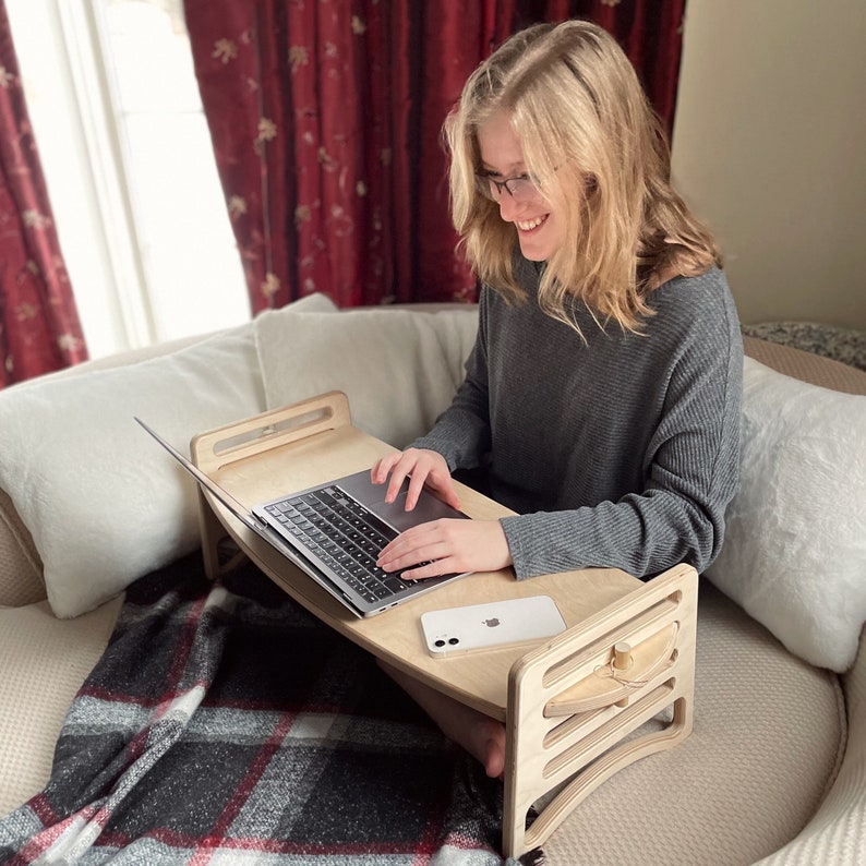 Large Adjustable lap desk being used by a woman sitting on a couch with two legs crossed. The desk is supporting a laptop and phone. We can clearly see the simple assembly mechanism with slots and dowel. Great gift for students