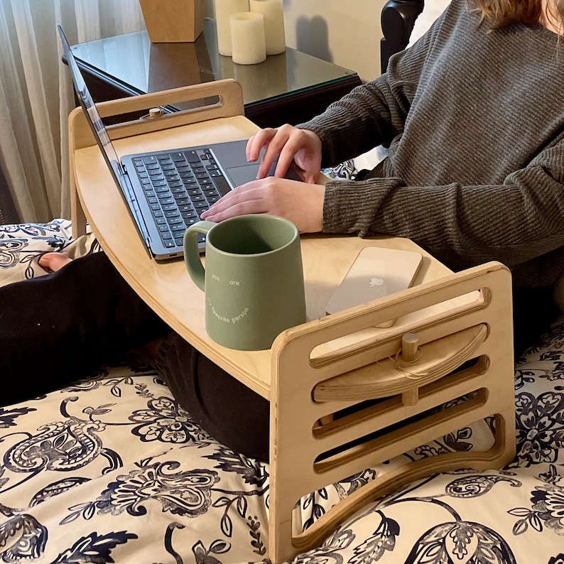 Large Adjustable lap desk being used by a woman sitting on a bed with one leg crossed. The desk is supporting a laptop, mug and phone. We can clearly see the simple assembly mechanism with slots and dowel. Great gift idea for a student dorm.