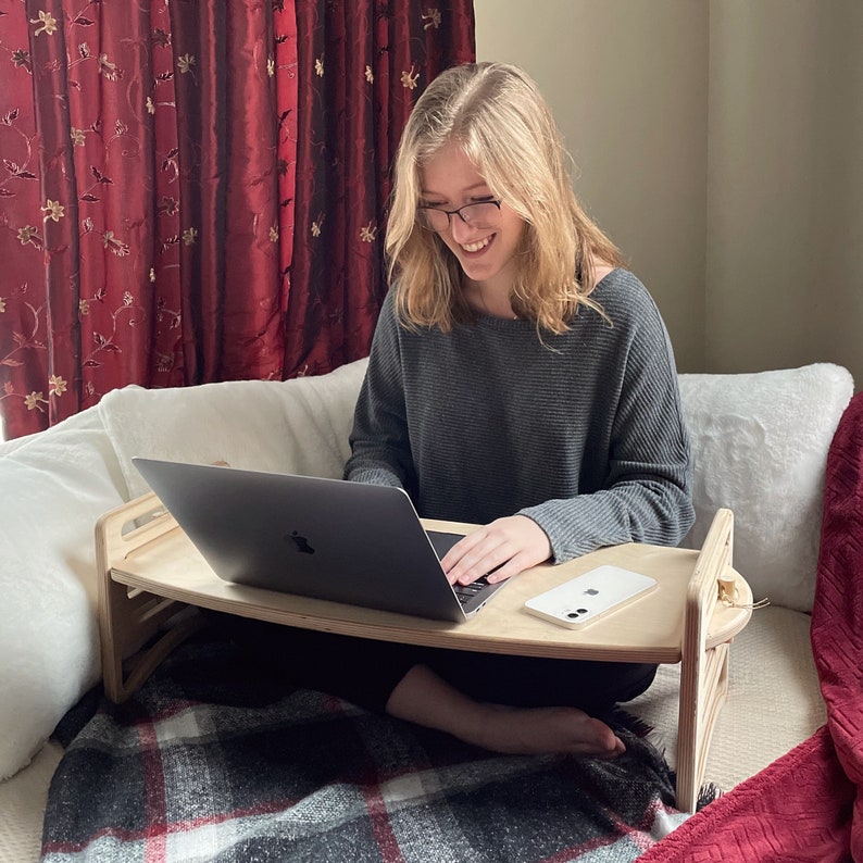 Large Adjustable lap desk being used by a woman sitting on a bed with one leg crossed. The desk is supporting a laptop, mug and phone. We can clearly see the simple assembly mechanism with slots and dowel.