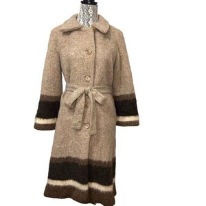 Peut inclure: Un long manteau en tricot beige avec des rayures brunes et blanches aux poignets et à l'ourlet. Le manteau a une ceinture et une fermeture à boutons.