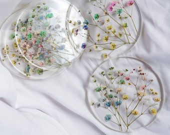 Puede incluir: Cuatro posavasos redondos y transparentes con flores secas de colores incrustadas en resina. Las flores incluyen tonos de rosa, azul, amarillo y verde. Los posavasos están colocados sobre una superficie blanca y texturizada.