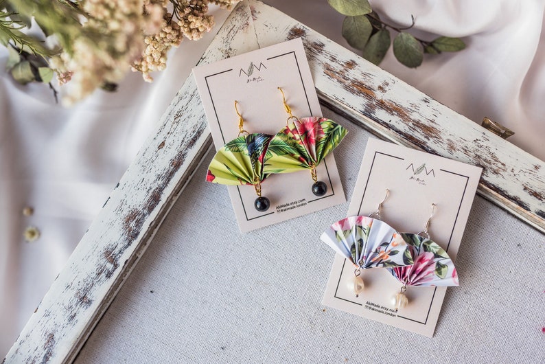 K&ouml;nnte beinhalten: Zwei Paar handgefertigte Origami-F&auml;cher-Ohrringe. Ein Paar zeigt ein gr&uuml;nes und wei&szlig;es Blumenmuster mit schwarzen Perlen. Das andere Paar zeigt ein rosa und wei&szlig;es Blumenmuster mit wei&szlig;en Perlen. Beide Paare werden auf wei&szlig;en Karten mit dem Text "Handgefertigt mit Liebe" und "@handmade_london" angezeigt.