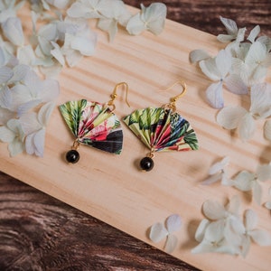 K&ouml;nnte beinhalten: Ein Paar Ohrringe mit einem floralen F&auml;cherdesign und schwarzen Perlenakzenten. Die Ohrringe liegen auf einer Holzoberfl&auml;che mit wei&szlig;en Bl&uuml;tenbl&auml;ttern.