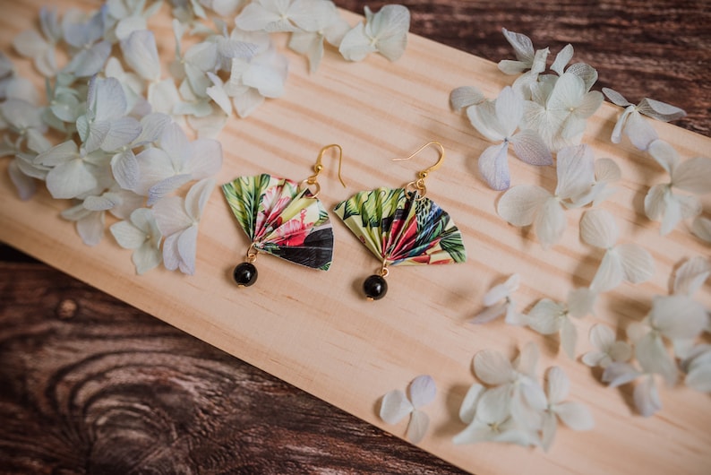 K&ouml;nnte beinhalten: Ein Paar Ohrringe mit einem floralen F&auml;cherdesign und schwarzen Perlenakzenten. Die Ohrringe liegen auf einer Holzoberfl&auml;che mit wei&szlig;en Bl&uuml;tenbl&auml;ttern.