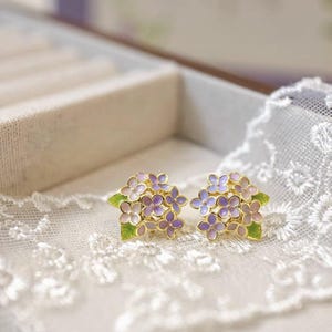 Könnte beinhalten: Ein Paar goldfarbene Ohrstecker mit Trauben aus lila und rosa Blüten mit grünen Blättern. Das florale Design ist detailliert und filigran, vor einem weißen Spitzengrund. Die Ohrringe sind ein Schmuckstück.