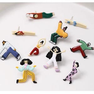 May include: A selection of quirky enamel pins featuring miniature figures in dynamic poses. The pins showcase a range of outfits in colours like red, green, yellow, and blue. Some figures have black hair and are in active poses, others are more relaxed.