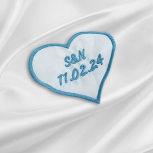 May include: A white heart-shaped patch with blue stitching. The patch has the text "S&N 11.02.24" embroidered in blue thread.