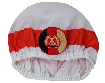 May include: A white shower cap with a red stripe and a circular patch featuring the flag of the German Democratic Republic. The patch has a black, red, and gold colour scheme and a stylised compass and hammer emblem.