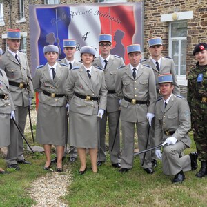 May include: A group of uniformed individuals, possibly military personnel, stand for a photo. They wear light grey jackets, skirts or trousers, and hats. Some hold batons. A French banner is visible in the background, with text in French.