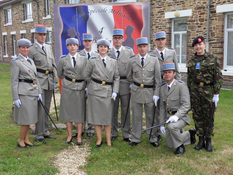 May include: A group of uniformed individuals, possibly military personnel, stand for a photo. They wear light grey jackets, skirts or trousers, and hats. Some hold batons. A French banner is visible in the background, with text in French.