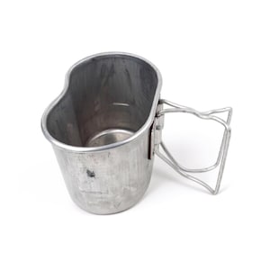 May include: A silver-colored metal canteen cup with a folding wire handle. The cup has an oval shape and a slightly textured surface. The handle is attached to the side of the cup with a hinge.