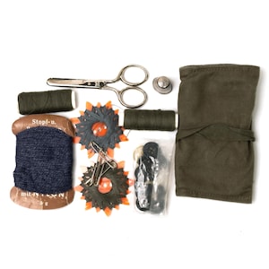 May include: A vintage sewing kit with various tools and supplies. The kit includes a pair of scissors, spools of thread, needles, buttons, a thimble, and a fabric case. The thread spools are dark green and navy blue. The kit is on a white background.