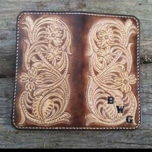 May include: A brown leather wallet with floral tooling. The wallet features a detailed embossed design of flowers and leaves. The initials "BWG" are visible on the lower right corner. The edges are stitched with white thread.