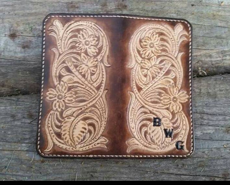 May include: A brown leather wallet with floral tooling. The wallet features a detailed embossed design of flowers and leaves. The initials "BWG" are visible on the lower right corner. The edges are stitched with white thread.